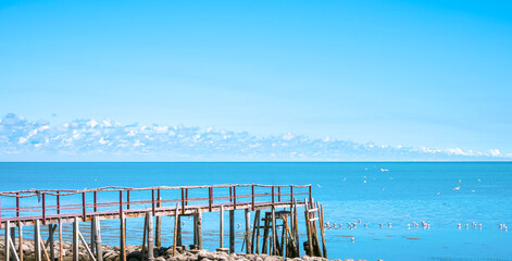 Wooden bridge extends into the sea in nature viewpoint area against blue summer sky background, perspective side view with copy space