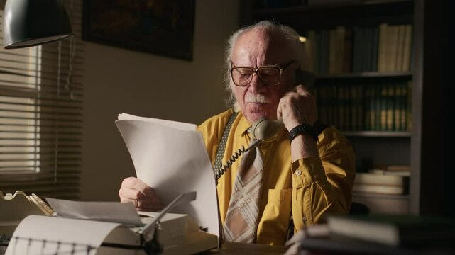 Medium shot of senior Caucasian male writer in yellow shirt holding typewritten script and talking to publisher on retro rotary phone working at desk in his vintage home study
