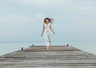 Asian woman jumping barefoot on wooden pier, calm sea horizon, dressed in white outfit under pale...