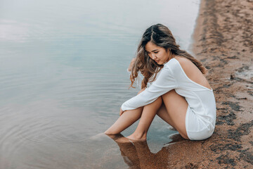 young Asian woman in white dress sitting on sandy lake shore gazing at water with thoughtful...