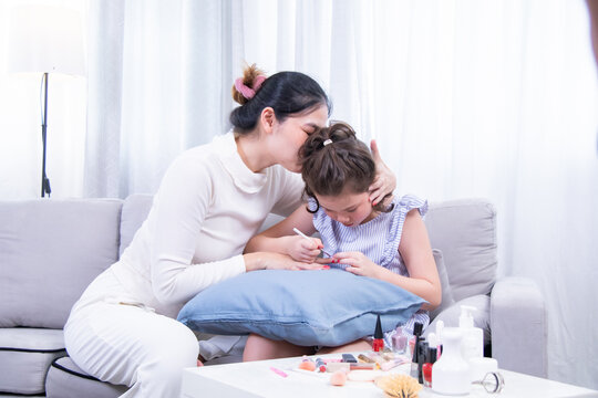 Kids, mother, bonding, beauty, lifestyle. A young girl focused on applying nail polish to her mother’s nails while sitting on a sofa. Sweet moment of care and creativity in their home beauty routine