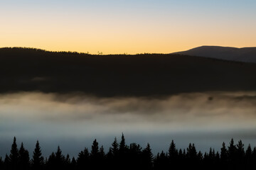 A serene mountain landscape at sunrise, with golden skies illuminating the horizon. A soft blanket of fog drifts over the dark forest below, while the snow-dusted peaks rise in the distance. 