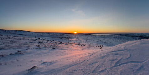 A breathtaking winter sunrise over a vast, snow-covered mountain plateau. The early morning sun casts a warm golden and pink glow across the untouched landscape.