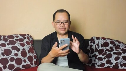 A middle-aged man wearing glasses is sitting on a couch and scrolling through a smartphone screen with focus - Powered by Adobe