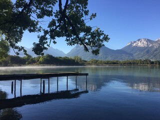 Serene lake landscape with misty waters reflecting distant mountains and a rustic wooden jetty...