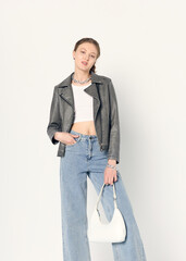 A happy woman with curly hair in a leather jacket with a bag in her hand poses against a light...