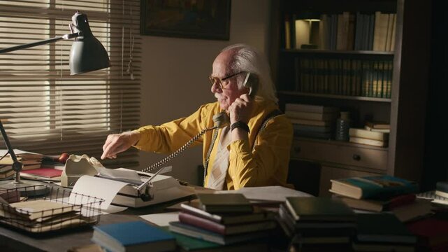 Medium side view shot of grey haired Caucasian male writer in yellow shirt sitting at desk in front of typewriter and lots of books and dialing number on retro rotary phone calling publisher