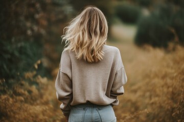 Obraz premium Blonde woman walking away through a field of tall grass wearing a sweater and jeans