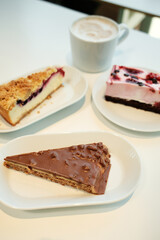 Three different cakes and one cappuccino arranged neatly on a white table for a perfect sweet break.