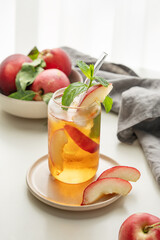 Ice tea with peach, mint in a glass on a white table near window with fresh fruits, napkin and sunlight.