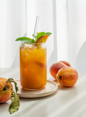 Ice tea with peach, mint in a glass on a white table near window with fresh fruits and shadows.