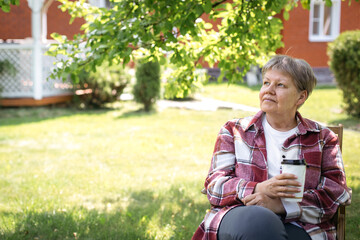 Elderly woman in red plaid shirt and white t-shirt sitting in garden with coffee cup on a sunny day, looking thoughtful, concept of reflection, theme of aging, lifestyle, relaxation, copyspace