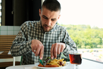 Close-up of young bearded man cutting schnitzel with fries and broccoli, enjoying lunch at a white...