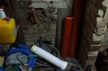 A disorganized basement area shows red and white pipes, blue hoses, and containers against a brick...