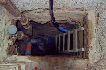 A work area shows a deep access point in the ground with a ladder, pipes, and a blue barrel,...
