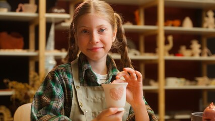 Positive smiling Caucasian teen little girl daughter child kid schoolgirl artist potter examining craft pottery clay vase cup admire creation enjoy hobby achievement looking at camera indoors workshop