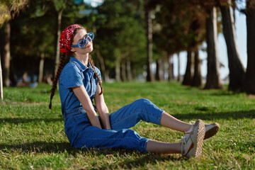 young woman sitting on grass wearing blue denim jumpsuit and red headscarf with sunglasses in sunny...