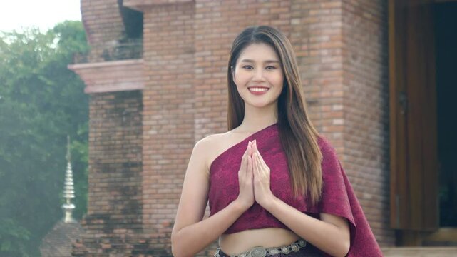 Smiling, beautiful young Asian woman wearing Thai traditional cloth greeting with Thai culture Sawasdee, welcome expression with temple in the background.