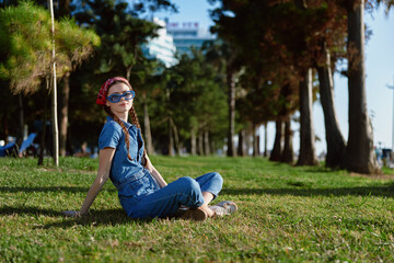 Fototapeta premium Lifestyle woman in modern boho-western denim street style sitting on grass outdoors wearing sunglasses and a headband, enjoying sunny weather with film color tones.