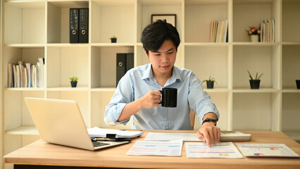 A business professional analyzing financial data and charts at a desk with a laptop, calculator, and documents in a bright office environment
