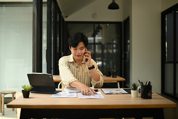 A Confident entrepreneur discussing projects remotely, surrounded by paperwork, a tablet on a...