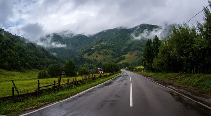 Naklejka premium The road leads to the mountains, with tree-lined mountain roads, cloud-shrouded mountains in front of and behind, and damp asphalt pavement. Forest after rain with misty mountain background.