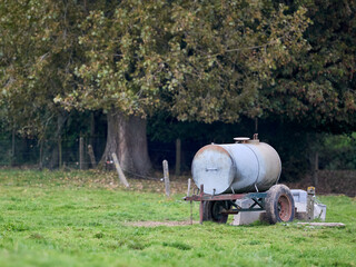 A weathered mobile water trough for livestock sits in a lush green pasture, with a large, leafy...