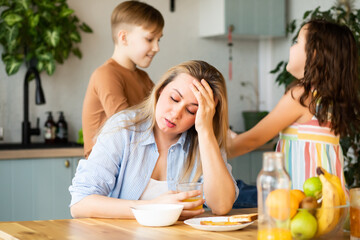 Young woman mom sitting on a kitchen table looking stressed suffers from headache because of noise...
