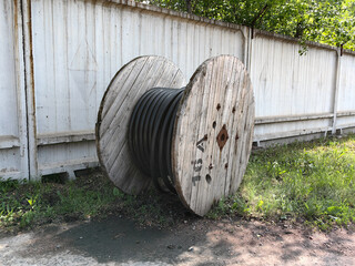 A large, wooden spool of cable rests in front of a weathered, white fence on a sunny day.