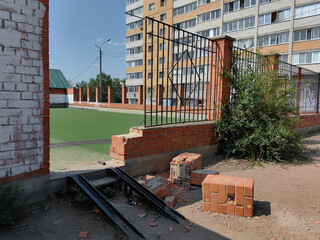A damaged brick wall and fence in front of an apartment building on a sunny day.