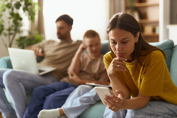 Mother text messaging on smart phone while relaxing next to her kids and husband on sofa in the...
