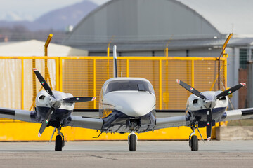 Front view of twin-engine piston propeller airplane on tarmac at small airfield with yellow fence backdrop
