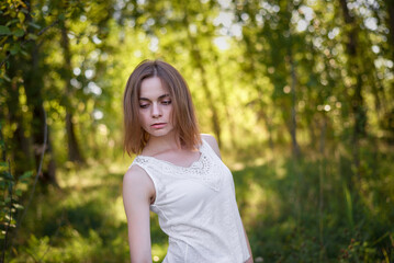 A beautiful woman stands in the forest on a summer day. She is wearing a white shirt and blue...