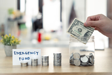A hand placing a dollar bill into a jar labeled 'Emergency Fund' with stacked coins nearby, representing saving money and financial preparedness.