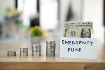 A hand placing a dollar bill into a jar labeled 'Emergency Fund' with stacked coins nearby, representing saving money and financial preparedness.