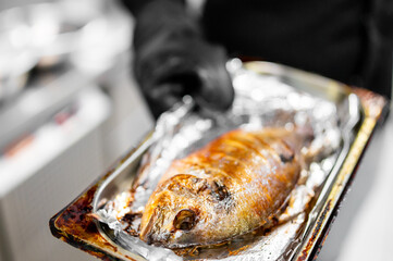 Golden brown baked fish on foil-covered tray, held with black glove. Close-up view of freshly...