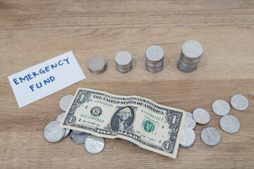 Clear jar marked emergency fund with stacked coins, folded dollar bills, and loose change on wooden table.