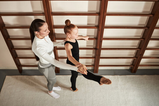 Caucasian young adult woman assisting Caucasian child girl practicing rhythmic gymnastics exercise at training facility, both standing near wooden wall bars, girl balancing on one leg