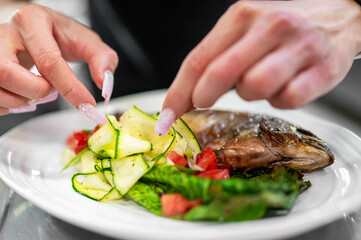 Woman plating grilled fish with zucchini ribbons, fresh salad, and herbs on a white plate. Close-up...