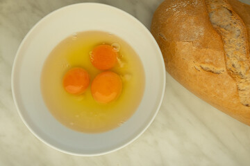 Cracked eggs in a white bowl on a light marble surface, with a loaf of bread placed beside it. 