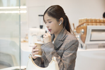 Young woman in plaid blazer enjoying coffee in modern cafe, wearing wireless earphones and holding...