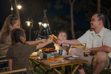 A family of four is sitting around a table with pizza and drinks. A man is holding a pizza slice and giving it to a child