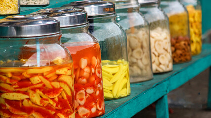 A variety of traditional Indonesian pickled fruit snacks are displayed in glass jars and baskets at local market stalls, displaying a vibrant and nostalgic street food scene.