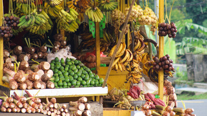 A vibrant tropical fruit and vegetable stall displays a colorful variety of fresh produce, including bananas, avocados, taro roots, and other local fruits. 