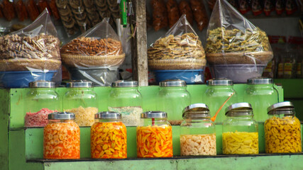 A variety of traditional Indonesian pickled fruit snacks are displayed in glass jars and baskets at local market stalls, displaying a vibrant and nostalgic street food scene.