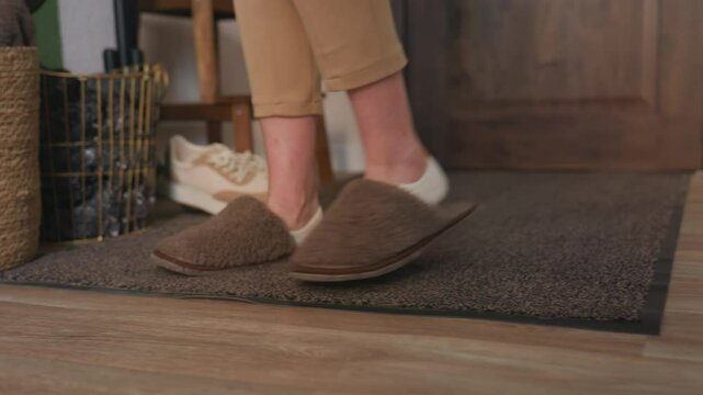 Leg view of individual wearing brown crocs walking onto foot mat, removing footwear and placing it into nearby basket, with beige pants, white socks, and wooden interior setting in softly lit room