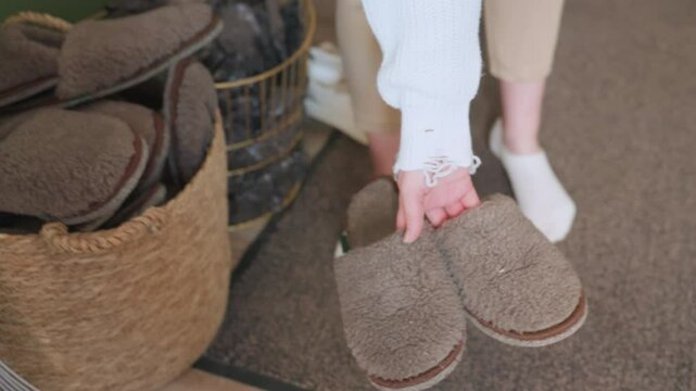 Lower view of guest selecting brown crocs from basket filled with slippers, wearing white socks and beige pants, preparing to put them on in indoor room with soft carpet, green wall, and wooden decor
