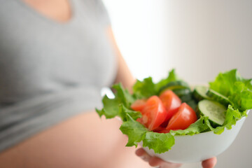 Expecting mother holding fresh vegetable salad while gently touching her belly. Healthy nutrition moment during pregnancy in cozy home setting.