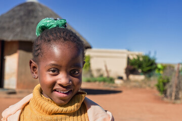 African village, single child girl with braids , standing in front of the house, wearing yellow...