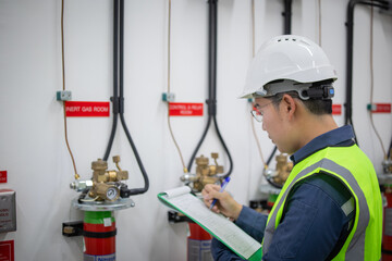 Worker wearing safety helmet and vest performs fire protection gas inspection while taking notes on clipboard in industrial setting ensuring safety and compliance